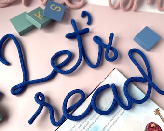 Puede incluir: Letras de lana azul que deletrean "Let's read" sobre una superficie rosa con un libro y bloques de madera.