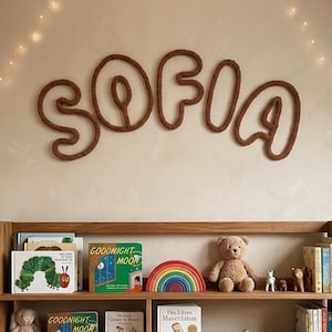 May include: A wooden bookshelf filled with children's books, toys, and a globe. The name "SOFIA" is spelled out in brown rope above the shelf. A teddy bear, rainbow, and other decorative items are on display.