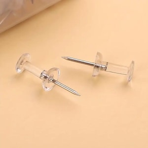 Puede incluir: Dos chinchetas transparentes con agujas de metal plateado. Cada chincheta tiene un cuerpo cilíndrico transparente con una cabeza circular y un disco circular más pequeño. Diseñadas para usar en un tablón de anuncios.