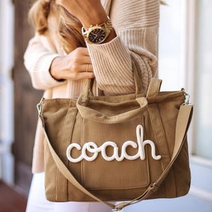 Può includere: Borsa tote in velluto a coste marrone con la scritta "coach" in corsivo bianco. La borsa ha una tracolla e due manici. Un orologio d'oro è visibile al polso.