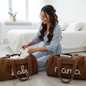 以下が含まれることがあります： 「baby」と「mama」のクリーム色の文字が入った、茶色のコーデュロイベビーバッグ2つ。バッグには茶色のハンドルが付いており、開いており、ベビー服やその他のアイテムが見えます。青いドレスを着た女性がアイテムを整理しています。
