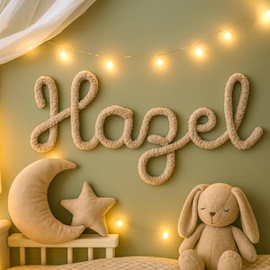 May include: A nursery wall decoration featuring the name "Hazel" in beige yarn, a crescent moon pillow, a star-shaped pillow, and a stuffed bunny. The items are set against a light green wall with string lights.