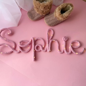 May include: A pink knitted wire sign spelling the name Sophie in cursive script, illuminated by small warm white fairy lights woven throughout the yarn. Two small brown cable-knit booties with faux fur trim are positioned above the name.