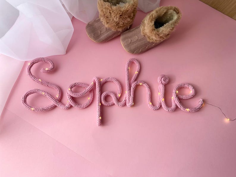 May include: A pink knitted wire sign spelling the name Sophie in cursive script, illuminated by small warm white fairy lights woven throughout the yarn. Two small brown cable-knit booties with faux fur trim are positioned above the name.