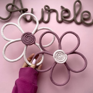 Peut inclure: Deux décorations florales faites à la main, une blanche et une mauve, avec un centre mauve. Les fleurs sont faites de corde et tenues par une personne. Le mot "Aurele" est écrit au-dessus des fleurs en corde marron sur fond rose.