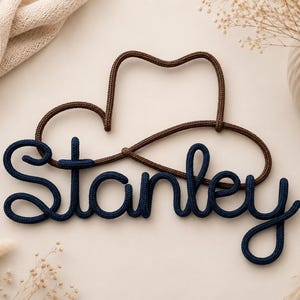 Op de afbeelding: Een wanddecoratie gemaakt van donkerblauw touw in de vorm van de naam Stanley, geplaatst voor een bruine touwomtrek van een cowboyhoed. De cursieve letters zijn verbonden in één doorlopende lijn, wat een gepersonaliseerd naambord vormt.