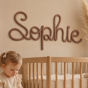 Op de afbeelding: Een naambord voor aan de muur met de naam Sophie, geschreven in een doorlopend, bruin, gebreid garenscript. De letters zijn gevormd uit een dik, getextureerd koord dat een zachte, afgeronde uitstraling op de muur creëert.