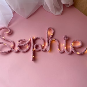 May include: A custom name sign spelling the word Sophie in cursive script, crafted from a dusty pink knitted yarn tube. The flexible structure is illuminated by a thin string of small, warm white LED fairy lights woven throughout the letters.