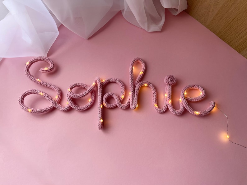 May include: A custom name sign spelling the word Sophie in cursive script, crafted from a dusty pink knitted yarn tube. The flexible structure is illuminated by a thin string of small, warm white LED fairy lights woven throughout the letters.