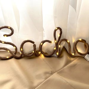 May include: A brown knitted wire sign spelling the name Sacha in cursive script, illuminated by small integrated warm white LED fairy lights. The letters are formed from a flexible cord with a visible knit texture and a small white battery pack.