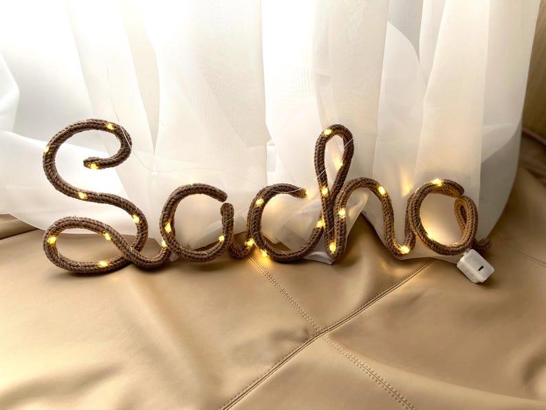 May include: A brown knitted wire sign spelling the name Sacha in cursive script, illuminated by small integrated warm white LED fairy lights. The letters are formed from a flexible cord with a visible knit texture and a small white battery pack.