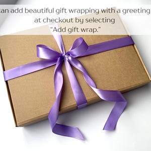 May include: A rectangular brown cardboard box tied with a smooth, shiny purple satin ribbon arranged in a bow. The text on the image reads: You can add beautiful gift wrapping with a greeting card at checkout by selecting Add gift wrap.