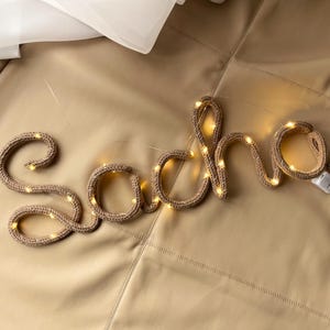 May include: A custom name sign spelling the word Sacha in cursive, constructed from a tan knitted fabric tube with integrated warm white LED fairy lights woven throughout the structure, featuring a small white battery pack attached at the end.