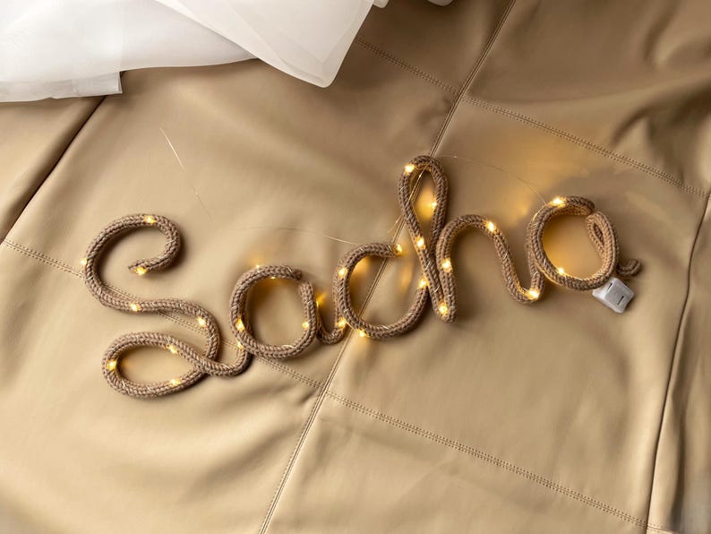 May include: A custom name sign spelling the word Sacha in cursive, constructed from a tan knitted fabric tube with integrated warm white LED fairy lights woven throughout the structure, featuring a small white battery pack attached at the end.