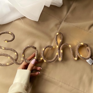 May include: A tan knitted yarn sign spelling the name Sacha in cursive script, featuring integrated warm white LED fairy lights throughout the letters and a small white battery pack attached to the end of the final letter.