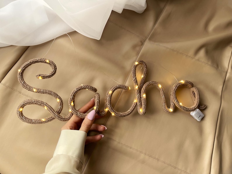 May include: A tan knitted yarn sign spelling the name Sacha in cursive script, featuring integrated warm white LED fairy lights throughout the letters and a small white battery pack attached to the end of the final letter.