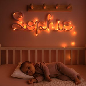 May include: A custom name sign spelling Sophie in cursive script, illuminated by warm white string lights. The sign is mounted on a wall above a white crib. The letters are formed by a flexible cord wrapped with glowing lights, creating a soft ambient glow.