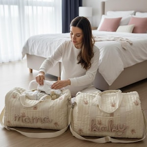 May include: Two cream-colored quilted baby bags with the words "mama" and "baby" embroidered in pink script. The bags have bunny face appliqués and are sitting on a wooden floor. A person is sitting between the bags.
