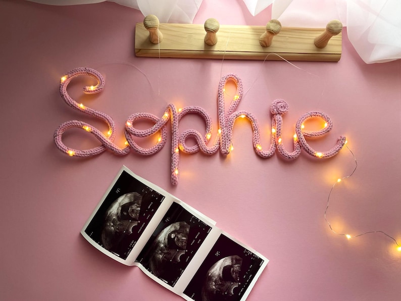 May include: A light pink knitted wire sign spelling the name Sophie, illuminated by a thin string of warm white LED fairy lights. Below the sign is a black and white ultrasound scan showing three distinct fetal images on a single paper strip.