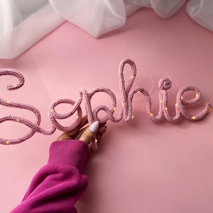May include: A custom name sign spelling the word Sophie in cursive script, crafted from pink knitted yarn wrapped around a flexible wire frame, featuring integrated warm white fairy lights woven throughout the letters for a soft glowing effect.
