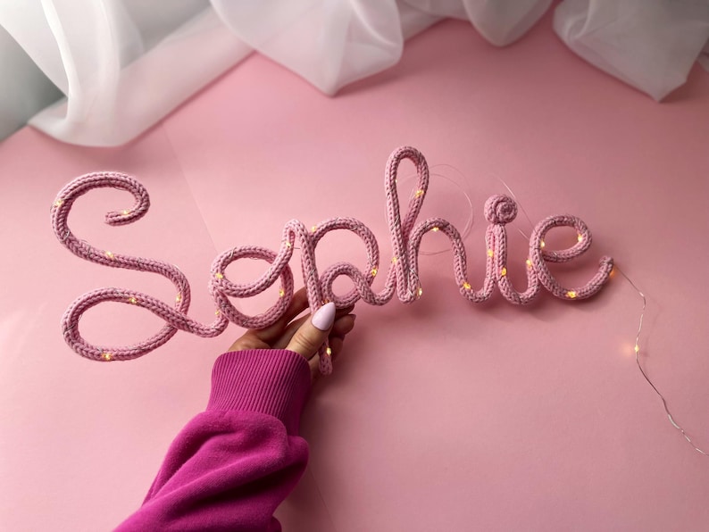 May include: A custom name sign spelling the word Sophie in cursive script, crafted from pink knitted yarn wrapped around a flexible wire frame, featuring integrated warm white fairy lights woven throughout the letters for a soft glowing effect.