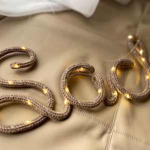 May include: A cursive word spelled out in a tan, knitted fabric tube with a flexible internal wire. The knitted structure is intertwined with a thin string of small, warm white LED lights that illuminate the tan yarn texture throughout the letters.