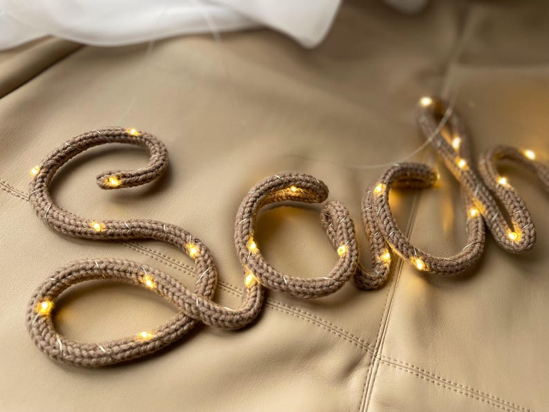 May include: A cursive word spelled out in a tan, knitted fabric tube with a flexible internal wire. The knitted structure is intertwined with a thin string of small, warm white LED lights that illuminate the tan yarn texture throughout the letters.