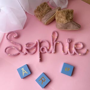 May include: A pink knitted wire sign spelling the name Sophie with integrated fairy lights, a pair of tan faux fur-lined booties, and three light blue square wooden blocks featuring the gold letters A, R, and a pair of baby footprints.
