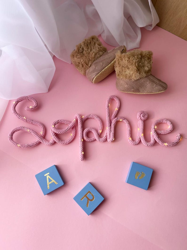 May include: A pink knitted wire sign spelling the name Sophie with integrated fairy lights, a pair of tan faux fur-lined booties, and three light blue square wooden blocks featuring the gold letters A, R, and a pair of baby footprints.