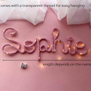 May include: A pink knitted wire sign spelling the name Sophie, illuminated by small warm white LED lights. The sign measures 5.25 to 6 inches in height and includes a transparent thread for hanging. A small battery pack is attached to the wire.