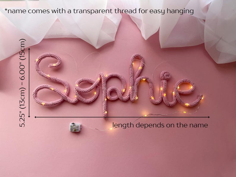 May include: A pink knitted wire sign spelling the name Sophie, illuminated by small warm white LED lights. The sign measures 5.25 to 6 inches in height and includes a transparent thread for hanging. A small battery pack is attached to the wire.