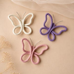 Könnte beinhalten: Drei schmetterlingsförmige Ornamente aus gestrickter Kordel in Creme, Lila und Rosa. Jeder Schmetterling hat zwei dünne, dunkle Drahtantennen, die aus dem Mittelkörper ragen, wobei die Flügel aus durchgehenden Schlaufen aus Stoff bestehen.