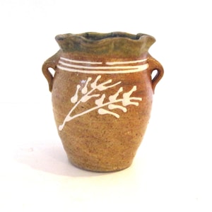 May include: A small, tan, speckled ceramic pot with two side handles and a ruffled, green-tinted rim. The exterior features a white, hand-painted botanical branch design and two horizontal white lines encircling the upper neck of the vessel.