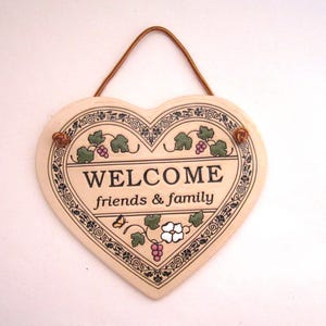 May include: Cream-coloured heart-shaped welcome sign with the words "WELCOME friends & family". It features a decorative border and grape vine accents. A brown rope is attached for hanging.