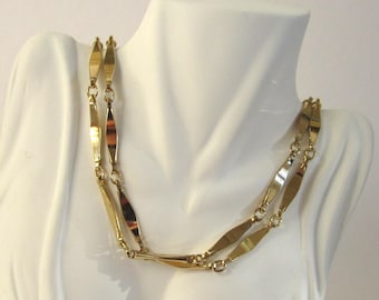 May include: A gold chain necklace with a double row of flat, elongated links. The necklace is displayed on a white mannequin bust.