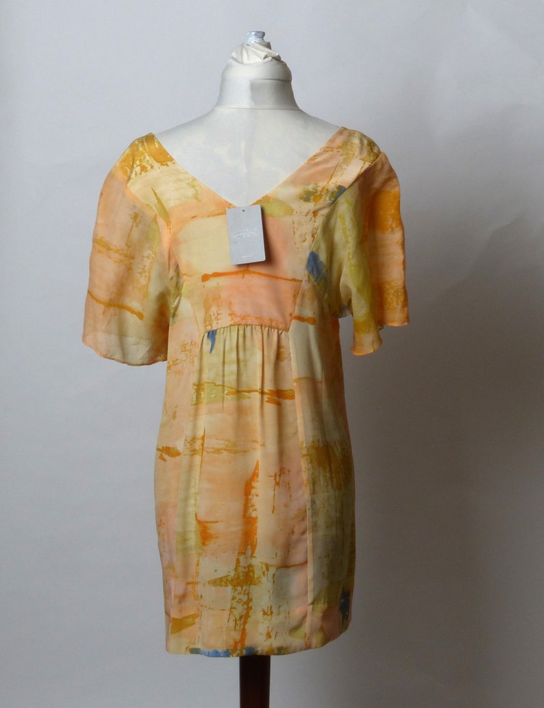 Puede incluir: Un vestido corto sin mangas con escote en V y estampado abstracto en tonos naranja, amarillo y azul. El vestido tiene una cintura fruncida y una silueta fluida.