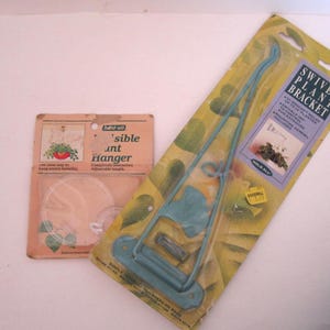 May include: Two vintage plant hangers are displayed. One is a clear plastic hanger with a printed image of a hanging plant. The other is a teal swivel plant bracket in its original packaging, with text that reads "Swivel Plant Bracket".