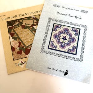 May include: Two instructional quilting pattern booklets. The left booklet is titled Heartfelt Table Runner with a heart-themed design. The right booklet is titled Floral Quilt Series Iris and Fans Quilt, featuring a purple and white floral quilt pattern.