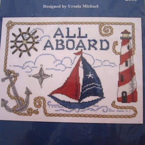 May include: Cross-stitch design with nautical elements. Features a sailboat with red and blue sails, a lighthouse, anchor, ship's wheel, and the words "ALL ABOARD." The design is framed with a rope border and set against a blue background.