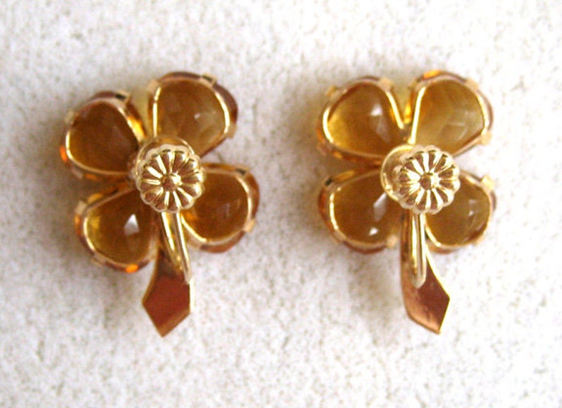 May include: A pair of gold tone earrings with four-leaf clover designs. Each clover is made of amber colored glass.