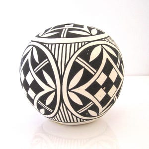 May include: A round, off-white ceramic vase with a black geometric pattern. The design includes repeating shapes of diamonds, leaves, and lines. The vase has a small opening at the top and is likely handmade.