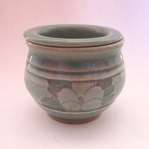 May include: A small, round, sage green ceramic butter crock with a glossy finish. The exterior features a hand-painted white flower design with dark green leaves. The vessel has a wide, rounded rim and a smooth, curved body shape.