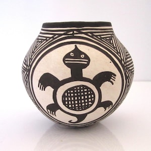 May include: A handcrafted ceramic pot with a rounded shape. The pot features a black and white design, including a central turtle motif and geometric patterns. The turtle has a grid-like shell. The rim is black.