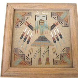 Può includere: Arte della sabbia incorniciata quadrata raffigurante una figura nativa americana con le braccia tese. L'opera presenta uno sfondo beige con la figura in tonalità di marrone, blu e rosso. La cornice è in legno chiaro.
