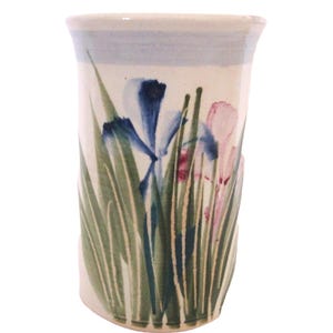 May include: A cylindrical ceramic vase with a cream-colored base featuring a hand-painted design of blue and pink iris flowers surrounded by tall green grass blades. The rim of the vase is accented with a soft light blue band around the top edge.
