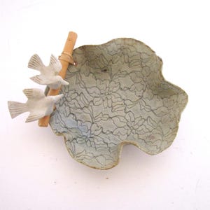 May include: A light green ceramic dish with an irregular, wavy edge and a dark grey floral lace pattern. Attached to the side is a light brown wooden stick featuring two small, white, sculpted birds perched on the surface of the ceramic bowl.