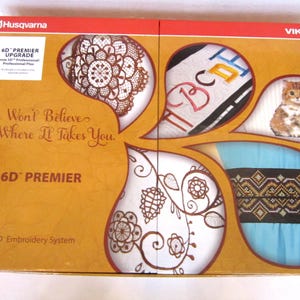 May include: An orange Husqvarna Viking 6D Premier embroidery system box. The box displays embroidery designs such as a lace pattern, embroidered letters, a cat, and a geometric pattern. The text on the box reads "You Won't Believe Where It Takes You."