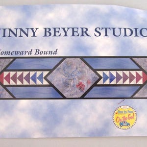 May include: A print with the text "JINNY BEYER STUDIO" and "Homeward Bound." The design features a central diamond shape with a bird and flag motif, framed by blue, purple, and red triangles. A circular logo is in the bottom right corner.