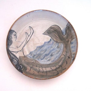 May include: A round ceramic plate with a hand-painted mermaid design. The mermaid has long dark hair and is depicted with her arms raised. The tail is brown and the water is blue. The plate has a brown rim.
