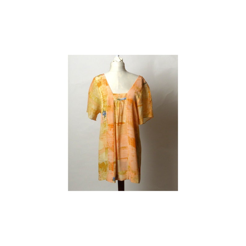 Puede incluir: Una blusa holgada de manga corta con cuello cuadrado. La blusa est&aacute; hecha de una tela ligera y presenta un estampado abstracto en tonos naranja, amarillo y azul.